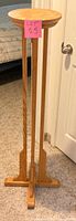 Tall wooden plant stand pedestal, showing full height and design of round top, vertical legs, and cross base, standing on carpet near a white door.