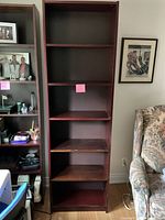 Front view of tall wooden shelving unit with five shelves, dark brown finish, positioned next to a floral upholstered chair and a framed artwork on the wall.
