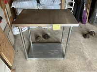 Rectangular stainless steel table with flat top and lower shelf, four cylindrical legs with reinforced feet, placed on concrete floor.