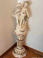 Full view of the plaster cherub figurine on its pedestal stand, showing overall shape, color, and decorative gold highlights.