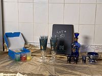 Blue and gold decanter with lid, matching tall liqueur glasses, small bowl, and box with colors of shot glasses shown alongside