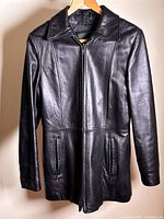Front view of black leather jacket hung on wooden hanger showing zip front and side zip pockets.