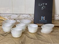 Several white opaque glass ramekins and onion soup bowls shown with white plastic lids stacked beside them on a marble surface.