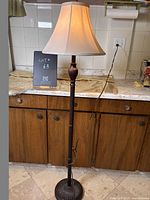 Full view of the wooden floor lamp against a kitchen backdrop, showing the beige fabric shade and carved wooden stand.