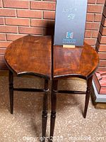 Front angle view of two vintage wooden half moon tables side by side on a brick background, showing top surface and legs.