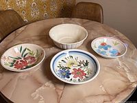 Four ceramic bowls placed on a round marble table, three with colorful floral designs and one plain with scalloped edges.