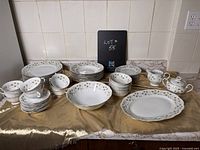 Full set of white porcelain Jarolina china with green leaf pattern, arranged with multiple plates in stacks, cups with saucers, bowls, serving platters, sugar bowl with lid, and creamer pitcher on a table covered with gold cloth.