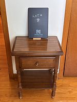 Front view photo of a vintage night stand showing the top, drawer, and shelf beneath, with a dark wood finish and turned legs.