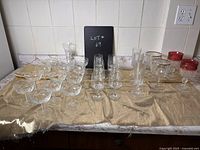 Clear glass parfait, wine, champagne, and beer goblets displayed on a beige cloth on a kitchen counter, with a black board showing lot number 69