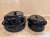 Two black speckled roasting pans shown with lids on a countertop; large pan on left, smaller pan on right