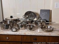 Photo shows assortment of stainless steel pots with lids, a vegetable steamer cone, colander, frying pan, and kettle arranged on kitchen countertop.