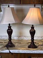 Two matching dark wood table lamps placed side by side on a marble surface with beige fabric shades and finials.