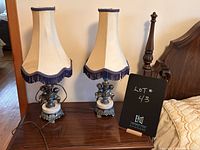 Two vintage cherub boudoir table lamps on wooden furniture with cream fabric shades trimmed in purple fringe, cherub base sculptures.