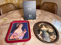 Photo of two vintage metal serving trays on table, Coca Cola rectangular tray on left, Pepsi Cola oval tray on right, with a black card labeled Lot #26 behind them.