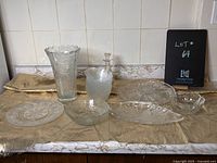 Photo shows nine pieces of vintage glassware on a beige cloth against a tiled wall, including a tall fluted vase, a cruet with stopper, various plates and bowls.