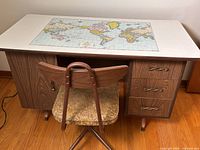 Front view of the vintage wooden desk with white top featuring a colorful world map design and three drawers on the right, accompanied by a mid-century style fabric upholstered chair with metal frame.