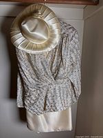 Cream satin pleated hat with bow atop printed wrap blouse hanging on a rod.