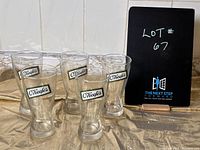 Five clear vintage O'Keefe's beer glasses arranged in a group with a black board reading 'Lot # 67' in the background.