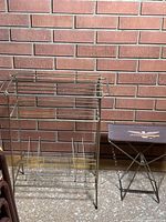 Full view of vintage metal vinyl LP record stand next to a foldable stool with fabric seat and metal frame against a brick wall.