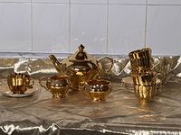 Full view of the entire Royal Winton gold-coloured tea set including teapot, cream and sugar bowl, and cups with saucers on a gold fabric backdrop.
