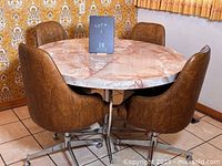 Round vintage kitchen table with marbled laminate top seen from an angle with four bucket chairs surrounding it.