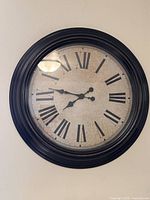 Front view of round plastic battery operated wall clock with black frame and Roman numerals on cream face.