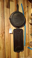 Two cast iron pans hung on wood wall, one round No. 10 with handle above a rectangular cast iron piece.