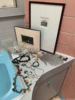 Photo of countertop surface with multiple costume jewelry pieces and two framed photos leaning against wall tiles