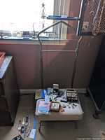 Brass look metal dressing stand next to a window with various items on a nearby table (not included). Stand features a tubular metal frame with horizontal and angled support bars.