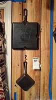 Two cast iron skillets hanging on wood plank: larger Colonial Breakfast Skillet #666 above smaller Square Egg Skillet #129 with tag visible