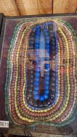 Photo showing all the beaded necklaces arranged in the wooden display case with glass covering, featuring blue, green, yellow, and brown beads.