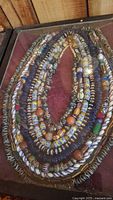 Overview of multiple Indigenous American glass and ceramic beaded necklaces arranged in a wooden display case with glass top, showing diverse intricate bead patterns and colors.