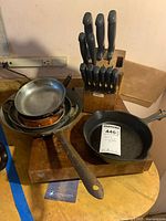 Wide angle photo showing Griswold cast iron pan, stacked copper pans, knife block filled with knives, and wooden cutting board on yellow table.