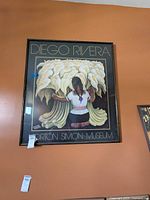 Framed Diego Rivera poster showing a person kneeling by calla lilies, hung on orange wall, with lot tag visible.