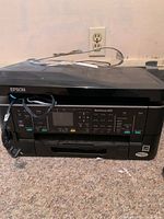 Front view of black Epson WorkForce 630 printer showing control panel with display and buttons for copy, fax, scan, and print functions. Power cable is visible.