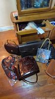 Photo showing multiple bags including a dark brown briefcase, purple handbag, a transparent floral tote, tan handbag, and a large embossed leather handbag on floor in front of drawers.