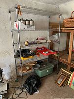 Front view of the metal shelving unit placed against a textured wall in an indoor space, showing five shelves with some miscellaneous items on them and nearby items on floor and adjacent wooden table.