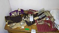 Overview of multiple silverware pieces rolled in cloth holders and spread on table, showing mixed utensils including knives, forks, spoons, and specialty items.