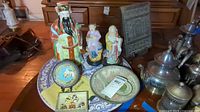 Wide shot showing all items on a table including ceramic figures, terracotta faces, large serving tray, metal plaque, and silver pots.