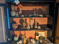 Wide shot showing shelves with vases, brass figurine bust, trinket boxes, souvenir seashells, glass containers, and framed art.