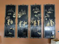 Set of four black lacquered Asian themed wall panels with detailed enameled figures and landscapes hanging on an orange wall.