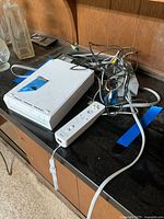 White Nintendo Wii console on a black countertop, blue tape on top, cables tangled behind, white Wii Remote in front.