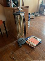 Electric wind chimes standing on wood floor next to a pile of folded fabrics or papers. Multiple silver metal tubes hanging from black circular frame held up by three black rods connected to a round base. Visible power adapter and wires attached.