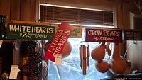 Photo of 5 painted wood signs hanging, showing text including White Hearts, Lanca Cigars, Crow Beads, and some wooden hanging items.