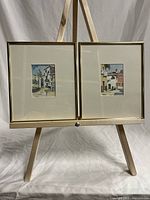 Pair of framed watercolour paintings displayed on wooden easel, gold-tone frames with cream mats, depicting architectural scenes of Italian villages.