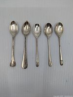 Five sterling silver teaspoons laid flat with their bowls facing upward, showing variation in handle designs and condition.