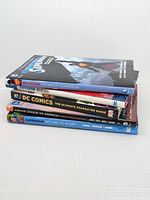 Five hardcover DC Comic books stacked horizontally. Titles visible from spine: Superman: The Men of Tomorrow, Superman: Reborn, DC Comics Ultimate Character Guide, Justice League of America, Superman: Return to Glory.