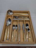 Complete cutlery set arranged in an expandable wooden drawer insert including forks, knives, spoons, and serving utensils with gold-plated accents.