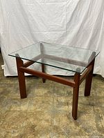 Front view of the teak wood coffee table with glass top showing the clear rectangular glass resting on a sturdy wood frame with straight legs and cross supports.