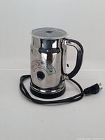 Front view of the Nespresso Aeroccino milk frother with power cord wrapped around base, showing stainless steel body, black handle, and instruction label.